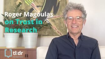 Roger Magoulas sits in a chair in his office with a painting and two plants on a cabinet behind his right shoulder